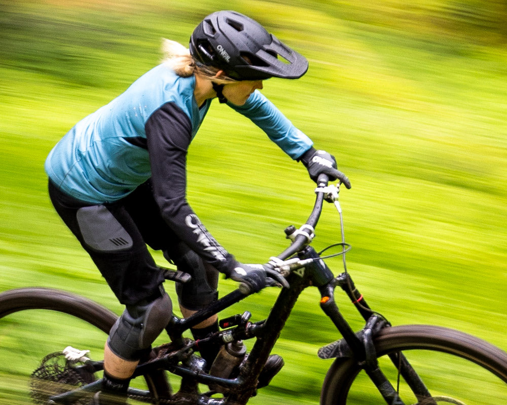 Eine Person mit Helm und Schutzausrüstung fährt mit einem Mountainbike schnell durch eine grüne, verschwommene Außenkulisse, was eine schnelle Bewegung suggeriert.