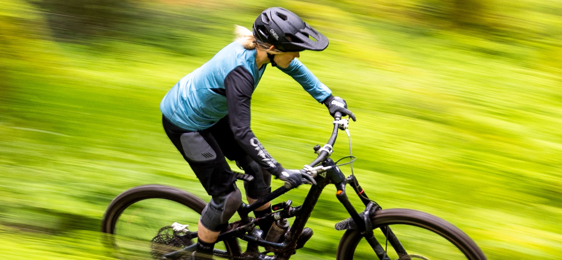 Eine Person mit einem blauen Langarmhemd, einer schwarzen Hose und einem Helm fährt schnell mit einem Mountainbike durch eine üppige, grüne, verschwommene Landschaft im Freien.