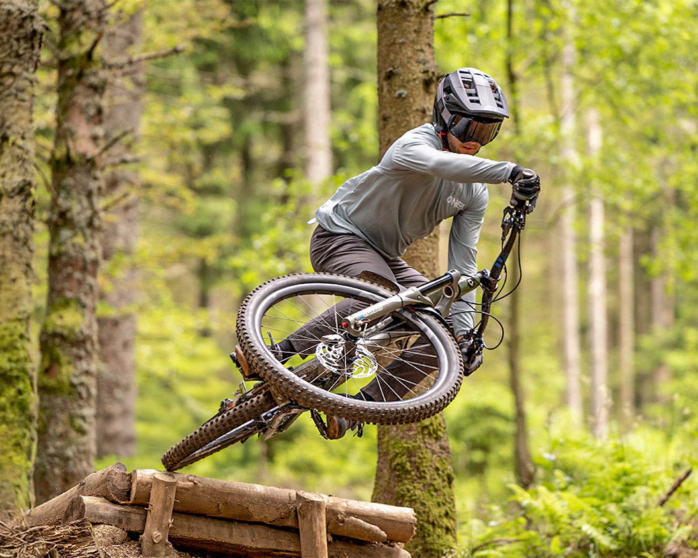 Ein Mountainbiker mit Helm und Schutzbrille springt von einer Holzrampe in einem üppigen, grünen Wald.