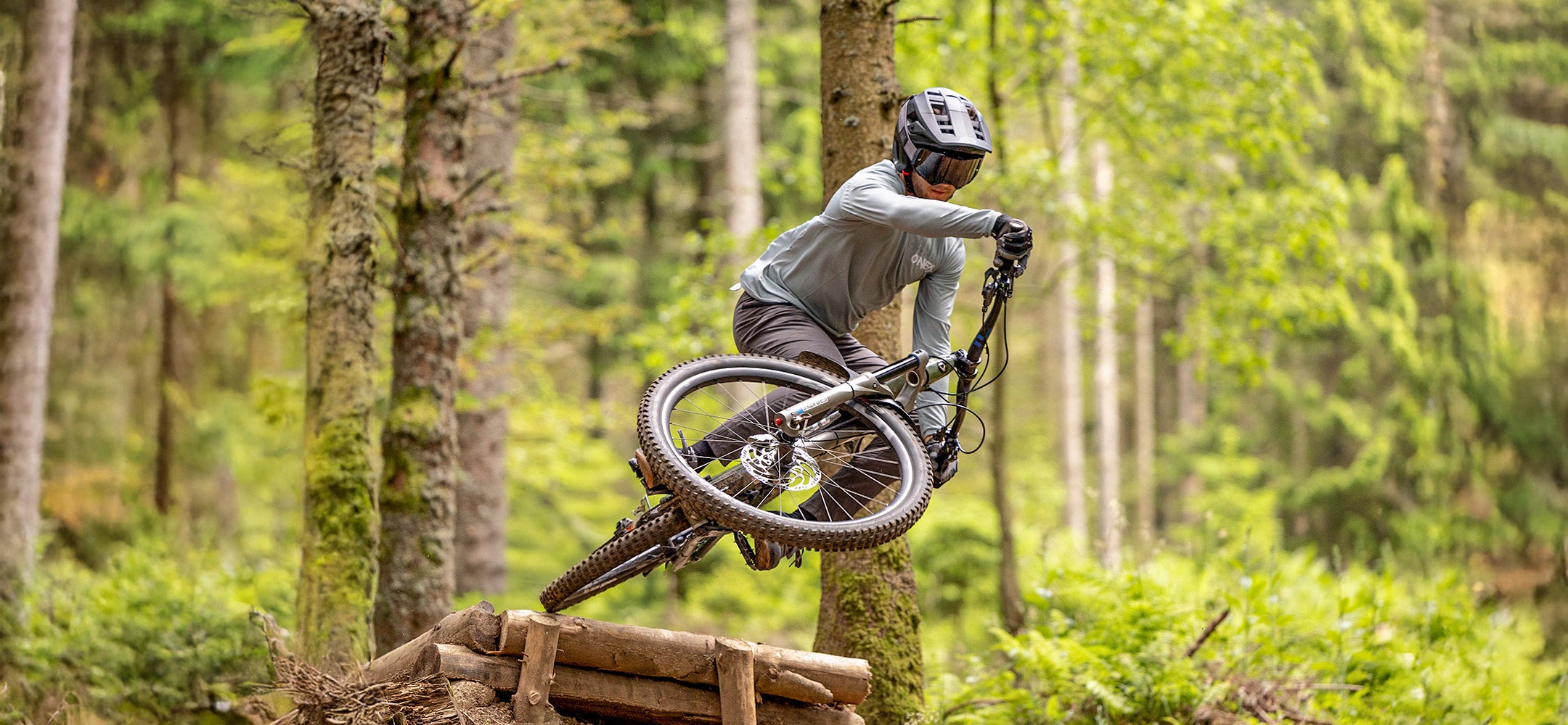 Ein Mountainbiker mit Helm und Schutzausrüstung fährt einen Feldweg in einem üppigen Wald hinunter und hebt dabei das Vorderrad von einer Holzrampe, während er von grünen Bäumen und Laub umgeben ist.