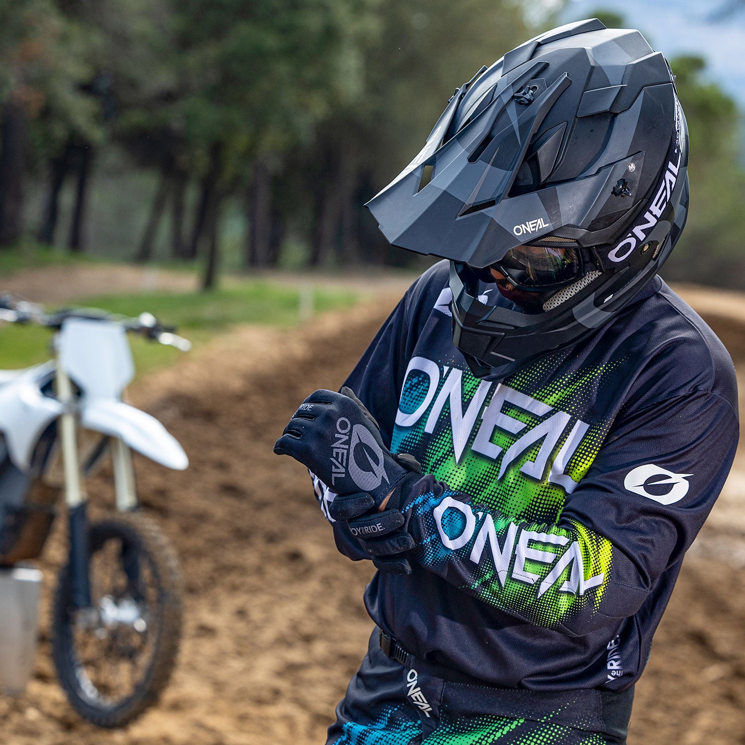 Eine Person mit schwarzem Motorradhelm und Handschuhen trägt eine schwarz-grüne O'Neal-Motorradkleidung. Sie richtet ihre Handschuhe, während sie auf einem Feldweg steht, im Hintergrund ist ein weißes Motorrad zu sehen. In der Ferne sind Bäume und Grün zu sehen.