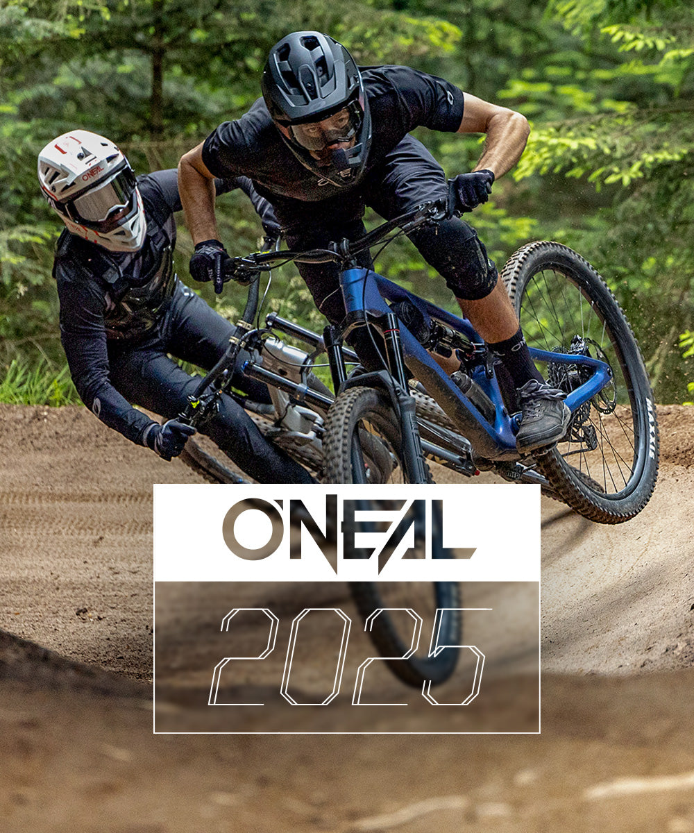 Zwei Mountainbiker mit Helmen und schwarzer Ausrüstung fahren dicht hintereinander um eine Kurve auf einem Feldweg im Wald. Am unteren Bildrand ist der Text „ONEAL 2025“ eingeblendet.