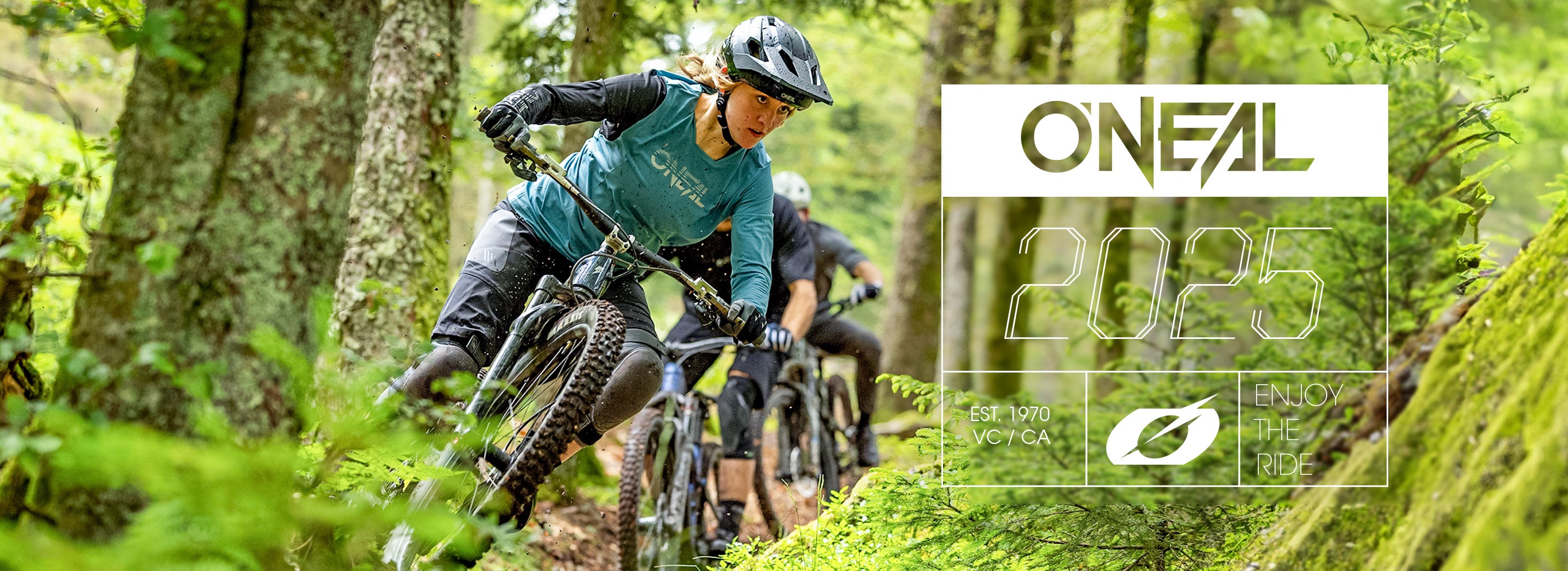Zwei Mountainbiker mit Helmen und O'Neal-Ausrüstung fahren zügig einen Waldweg entlang, der von üppigem Grün umgeben ist. Auf dem Bild steht „O'NEAL 2025“ und „Enjoy the Ride“.