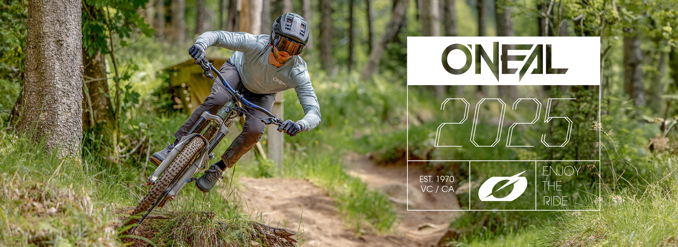 Ein Mountainbiker in Schutzkleidung fährt einen Trail durch einen grünen Wald. Der darüber gelegte Text lautet „ONEAL 2025“, „EST. 1970 VC/CA“ und „ENJOY THE RIDE“ mit einem Blattsymbol.