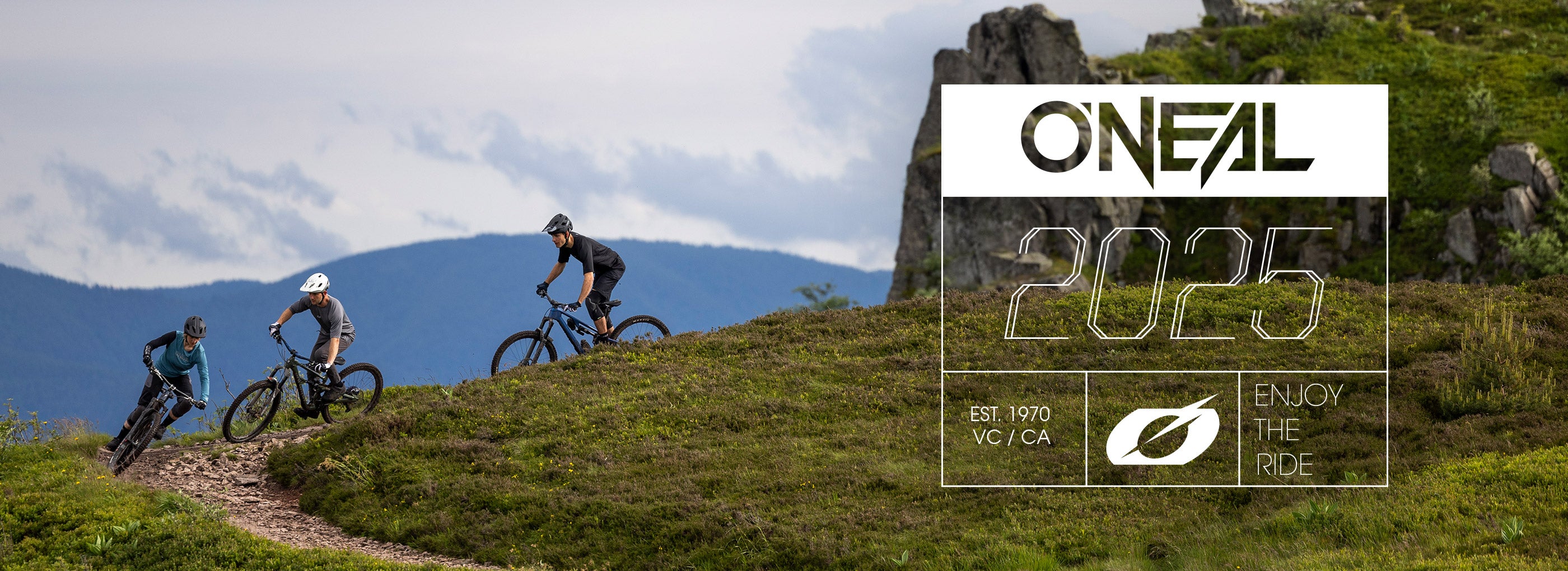 Drei Radfahrer fahren mit Mountainbikes auf einem grasbewachsenen Weg, der sich durch die Hügel schlängelt, mit Bergen im Hintergrund. Das Bild zeigt den Text „ONEAL 2025“ und den Slogan „Enjoy the Ride“.