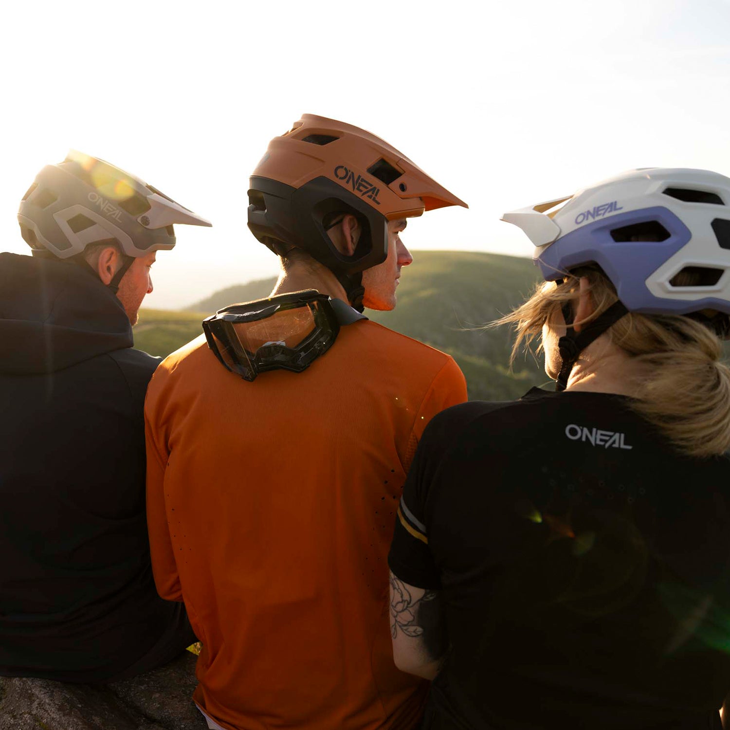 Drei Menschen mit Fahrradhelmen und Radsportausrüstung sitzen zusammen im Freien auf einem Felsen, den Blick von der Kamera abgewandt, und blicken auf eine malerische, sonnenbeschienene Landschaft mit grünen Hügeln im Hintergrund.