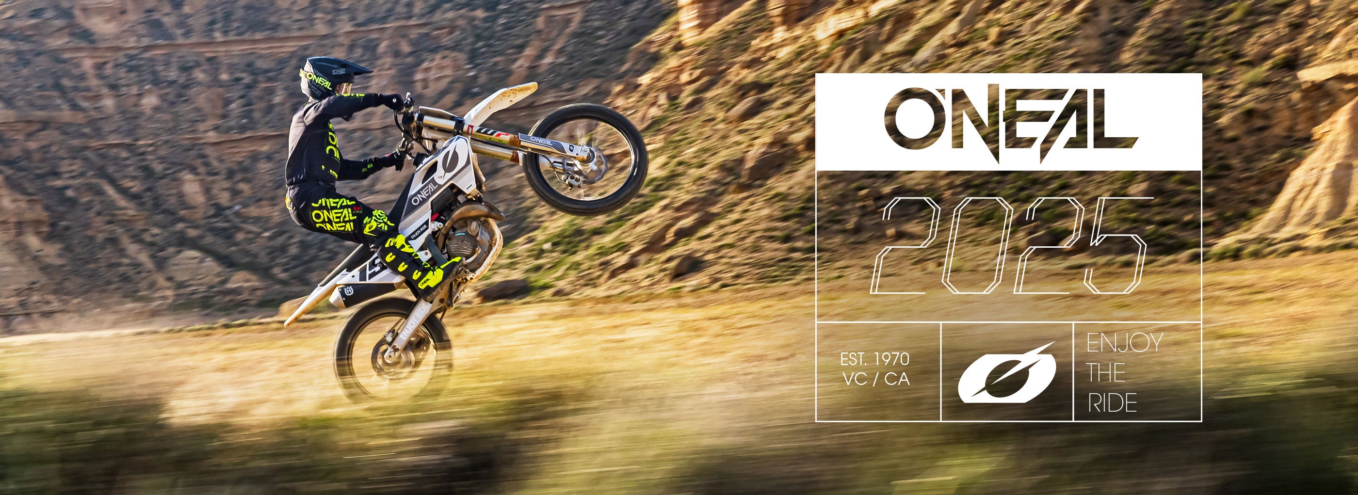 Eine Person in O'NEAL-Ausrüstung macht einen Wheelie auf einem Dirtbike in einer felsigen Landschaft. Der Overlay-Text lautet: „O'NEAL 2025, Est. 1970 YC/CA, Enjoy the Ride“ mit einem Blattsymbol.