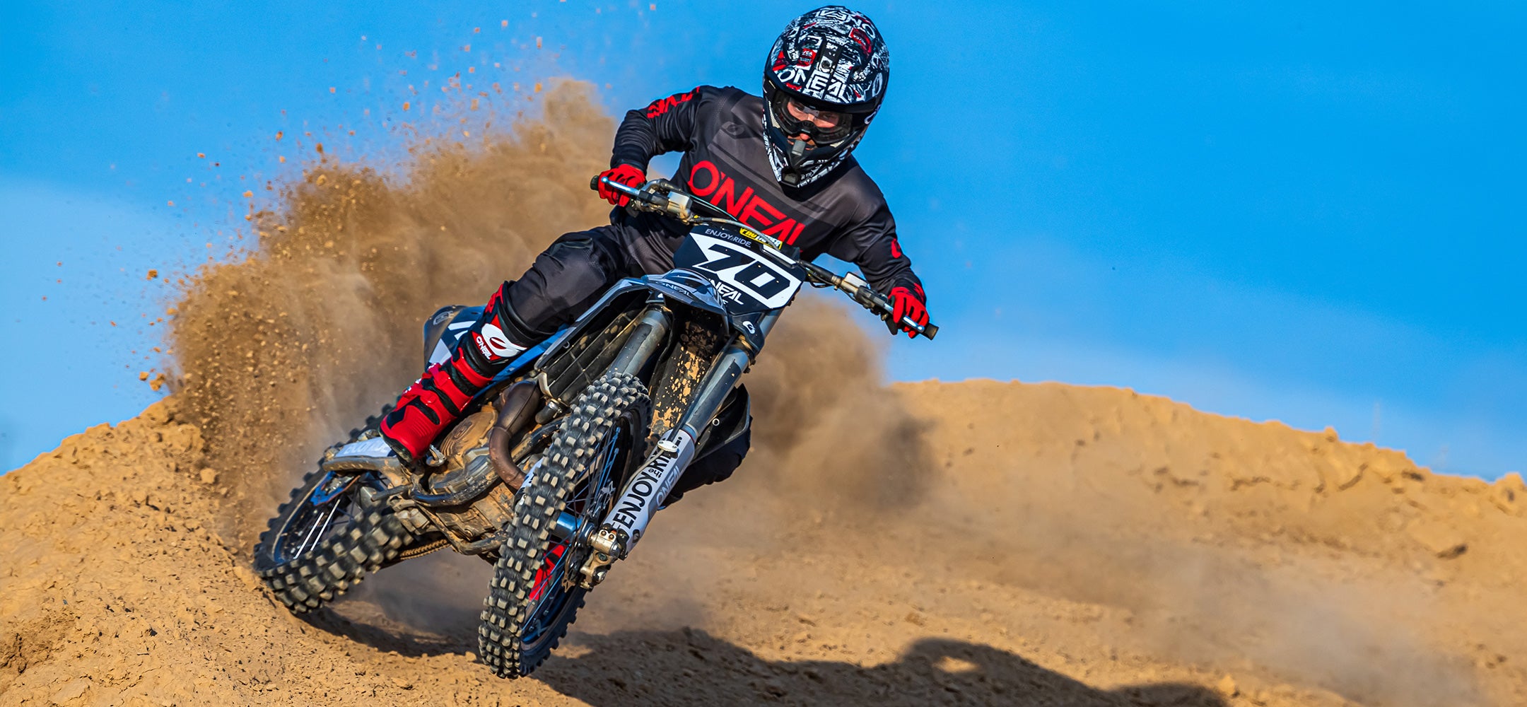 Ein Motocross-Fahrer in schwarz-roter Ausrüstung rast mit einem Dirtbike um eine sandige Kurve und wirbelt dabei vor einem klaren blauen Himmel eine große Sandwolke auf.