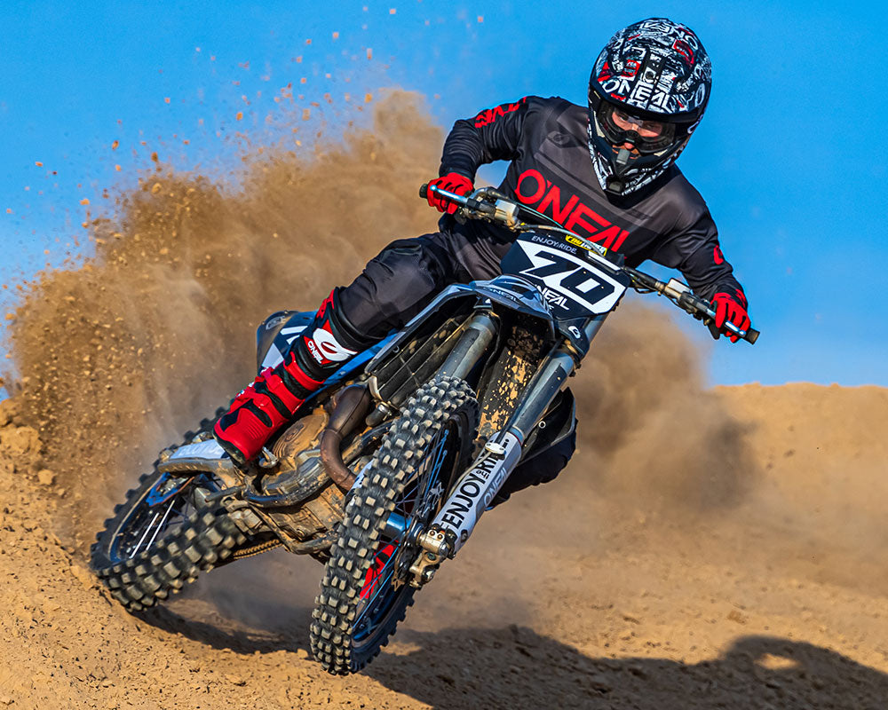 Ein Motocrossfahrer in voller Montur, einschließlich eines schwarzen Helms mit rot-weißen Mustern, fährt eine Sandpiste. Als er sich in eine Kurve legt, wirbelt hinter dem Hinterreifen des Motorrads Staub auf. Der Himmel ist klar und blau.