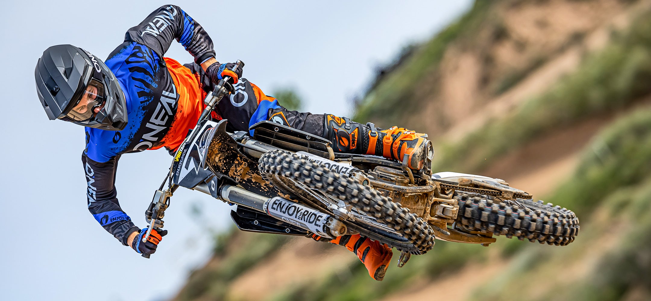 Ein Motocrossfahrer in blauer, oranger und schwarzer Ausrüstung führt auf einem Dirtbike einen Stunt in der Luft aus. Der Hintergrund zeigt eine verschwommene Feldwegstrecke mit etwas Grün, was die dynamische Bewegung und Geschwindigkeit der Szene betont.