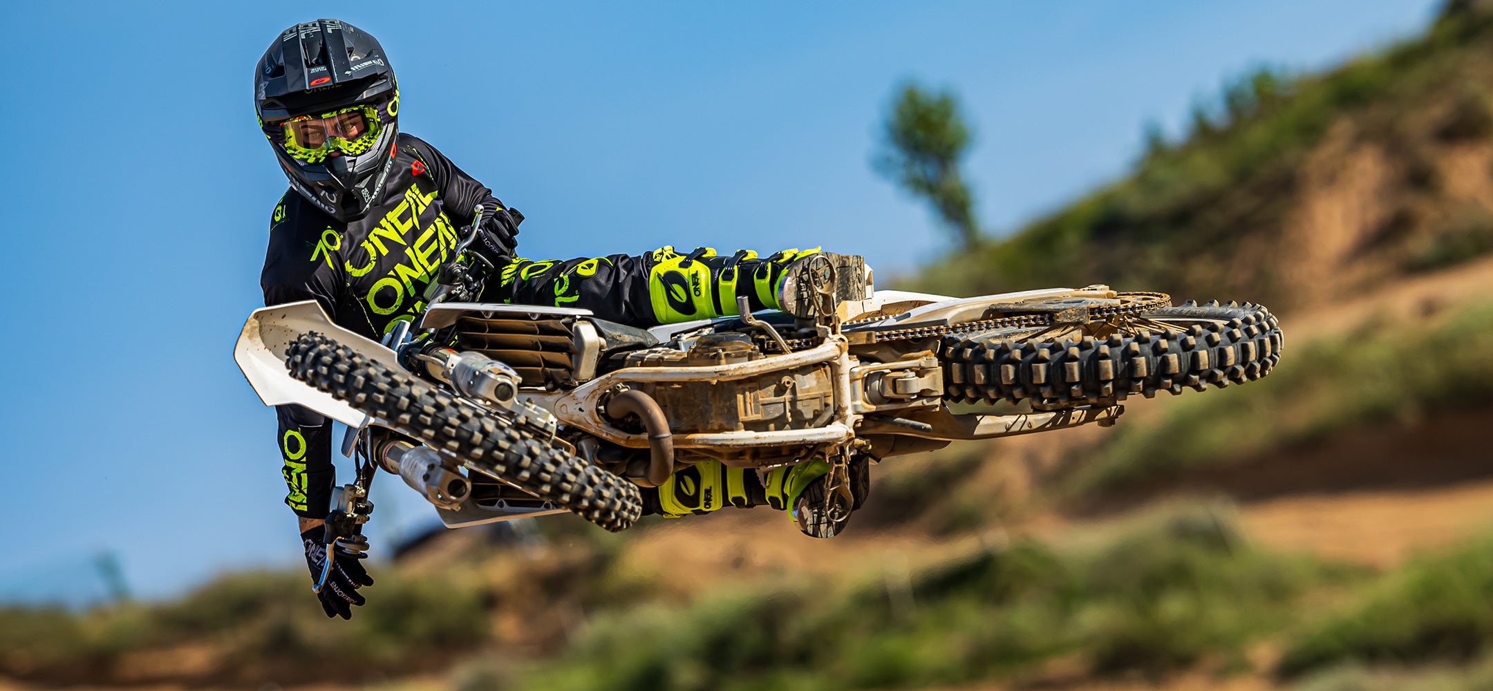 Ein Motocrossfahrer in schwarz-grünem Outfit und Helm führt vor einer unbefestigten Strecke und einem klaren blauen Himmel auf einem Dirtbike einen Flugtrick aus. Das Bike ist seitlich geneigt, sodass die Unterseite und die beeindruckende Kontrolle des Fahrers zu sehen sind.