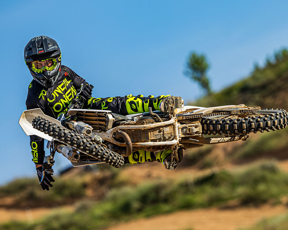 Ein Motocrossfahrer in schwarzer und neongrüner Ausrüstung führt auf einem Dirtbike einen Stunt in der Luft aus. Das Bike ist horizontal geneigt und der Fahrer streckt einen Arm aus. Der Hintergrund ist ein blauer Himmel und ein unscharfes natürliches Gelände.