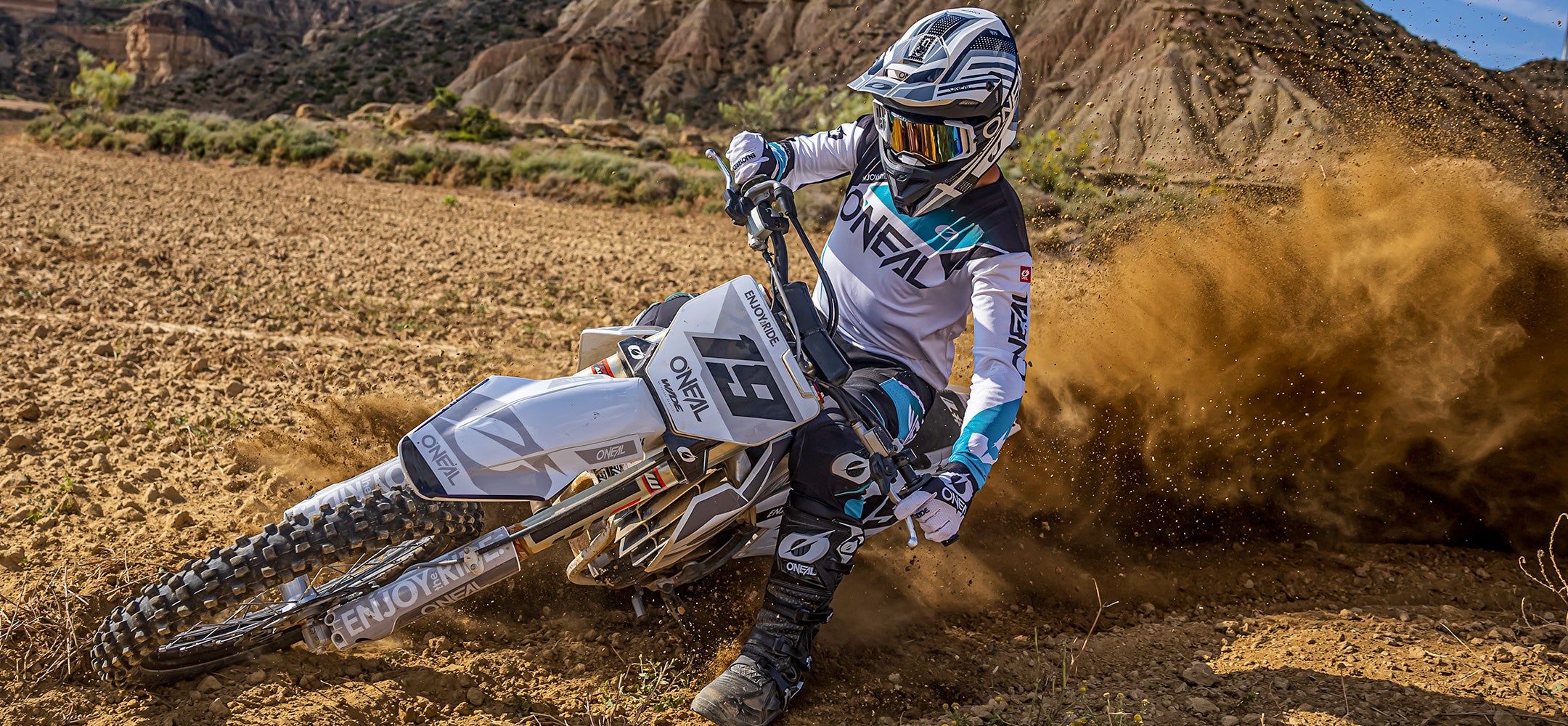 Ein Motocrossfahrer in weiß-blauem O'Neal-Outfit und Helm wirbelt eine Staubwolke auf, während er auf einem Feldweg in einer zerklüfteten, felsigen Landschaft eine scharfe Kurve fährt. Das Motocross-Bike ist angewinkelt und zeigt die intensive Action und Geschwindigkeit des Sports.