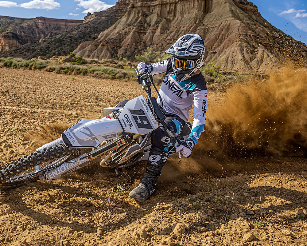 Ein Motocrossfahrer in voller Montur und mit Helm fährt mit einem Dirtbike durch ein sandiges, wüstenähnliches Gelände und wirbelt dabei eine Staubwolke auf. Im Hintergrund sind Felsformationen und ein klarer blauer Himmel mit ein paar Wolken zu sehen. Der Fahrer legt sich in eine scharfe Kurve.