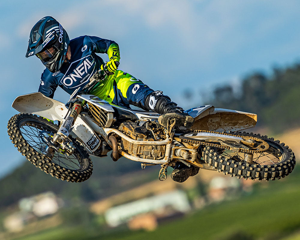 Ein Motocrossfahrer in O'Neal-Ausrüstung springt mit einem weißen Dirtbike über eine Dirtjump-Sprungfläche. Der Fahrer befindet sich in der Luft, mit den Rädern in der Luft, vor dem Hintergrund einer verschwommenen Landschaft mit Bäumen und Feldern.