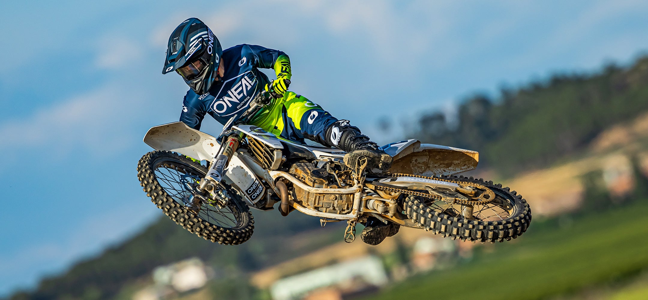Ein Motocrossfahrer in blau-grünem Outfit und Helm springt mit einem Dirtbike hoch in die Luft vor einem unscharfen natürlichen Hintergrund mit Bäumen und Gebäuden. Der Körper des Fahrers ist angewinkelt, was auf einen fortgeschrittenen Stunt hindeutet.