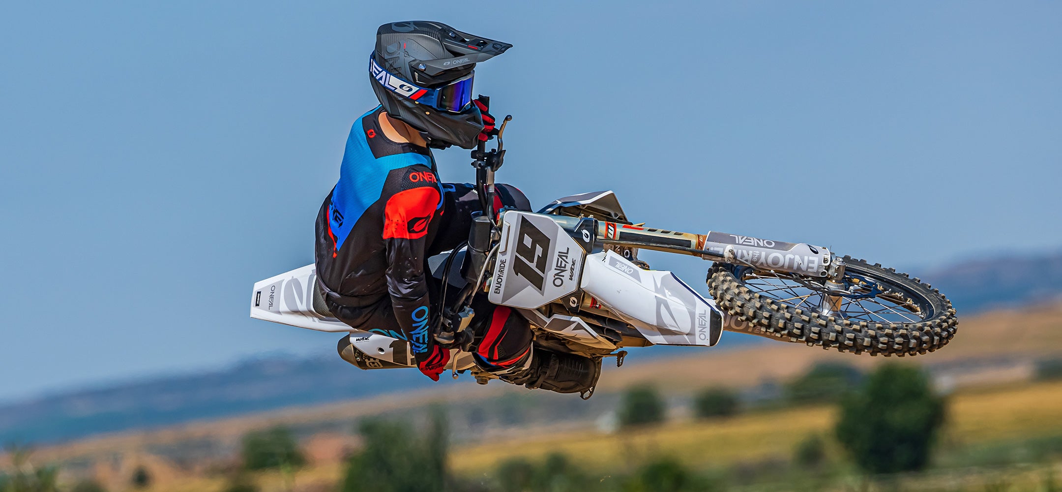 Ein Motocrossfahrer führt einen Trick in der Luft aus, bei dem er das Motorrad horizontal kippt. Der Fahrer trägt einen schwarzen Helm und blaue und rote Ausrüstung, und das Motorrad ist überwiegend weiß. Der Hintergrund zeigt eine Landschaft mit Gras und Bäumen, was auf eine Außenkulisse hindeutet.