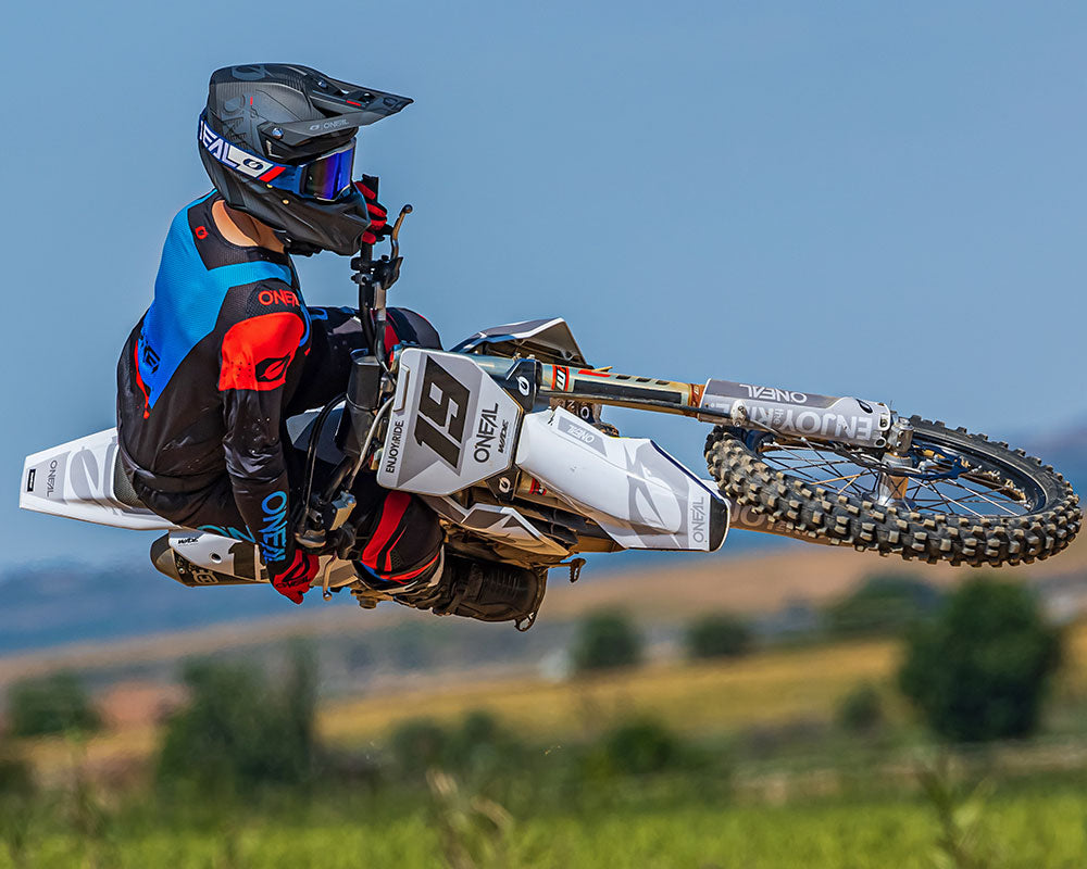Ein Motocrossfahrer vollführt in der Luft ein dynamisches Whip-Manöver auf einem weißen Dirtbike. Der Fahrer trägt ein rot-blau-schwarzes Outfit mit Helm und Schutzbrille. Im Hintergrund ist eine verschwommene Landschaft mit Feldern und Hügeln unter einem klaren blauen Himmel zu sehen.