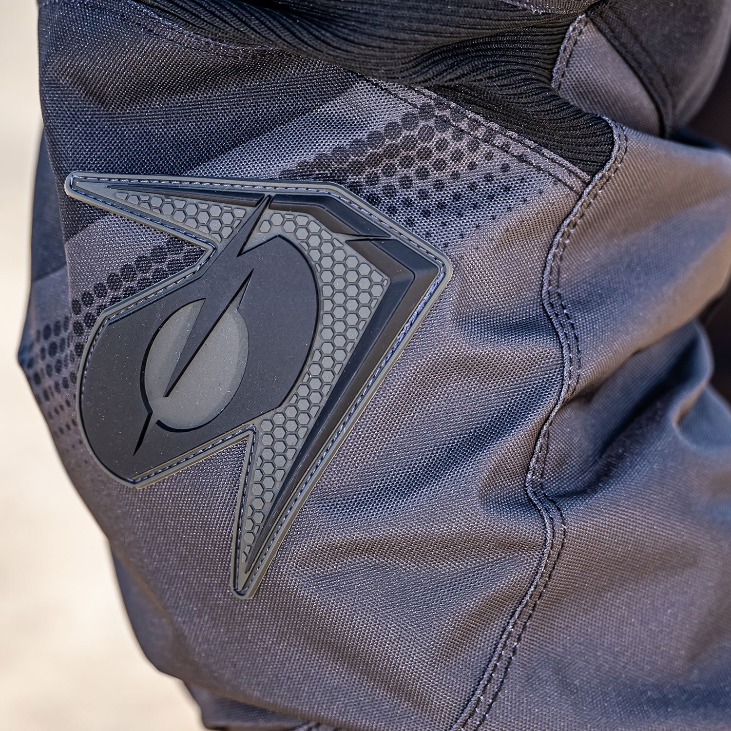 Nahaufnahme der Hose eines Motorradfahrers, mit einem detaillierten Aufnäher mit einem stilisierten Emblem. Die Hose besteht aus strapazierfähigem, strukturiertem Stoff, hauptsächlich in Grau und Schwarz, wobei das Emblem komplizierte Muster und eine halbreflektierende Oberfläche aufweist.