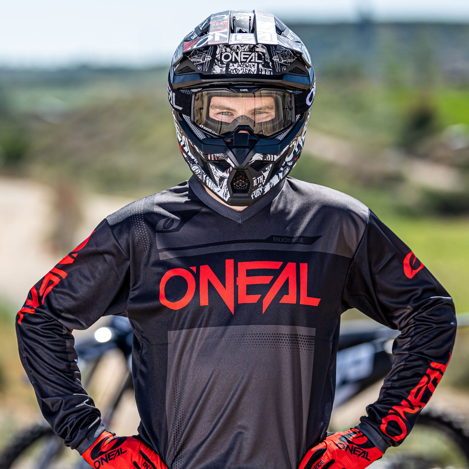 Eine Person in einem schwarz-roten Motocross-Outfit, einschließlich Helm mit Visier und Handschuhen, steht mit den Händen in den Hüften. Der Helm hat ein schwarz-weiß-graues Grafikdesign. Der Hintergrund zeigt eine verschwommene Außenkulisse mit Grün und einem Pfad.