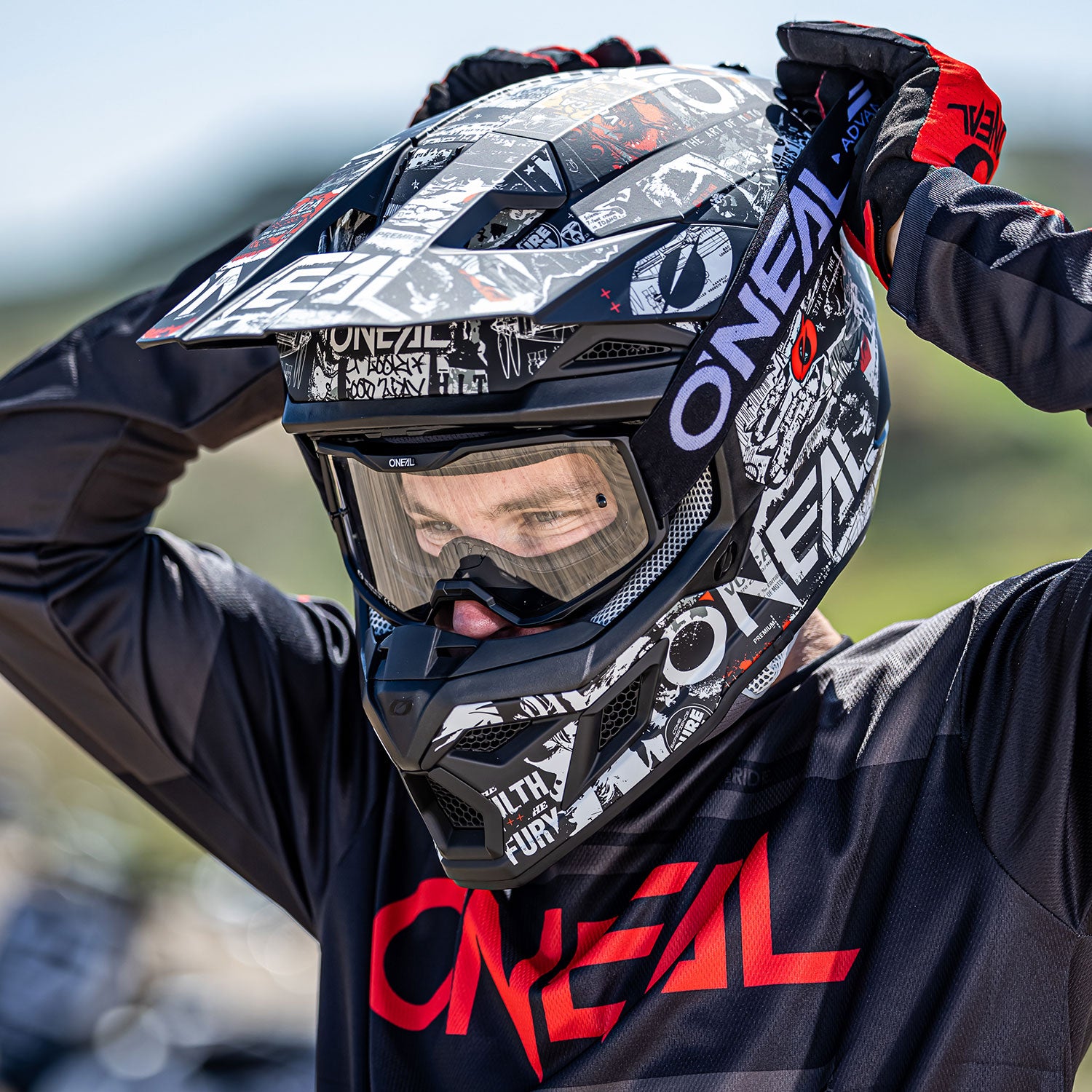 Eine Person mit einem schwarz-roten O'Neal-Motocrosshelm und passender Ausrüstung rückt ihren Helm zurecht. Der Helm weist verschiedene grafische Designs auf. Die Person ist im Freien, mit einem unscharfen Hintergrund, der eine Offroad- oder Motocross-Streckenumgebung suggeriert.