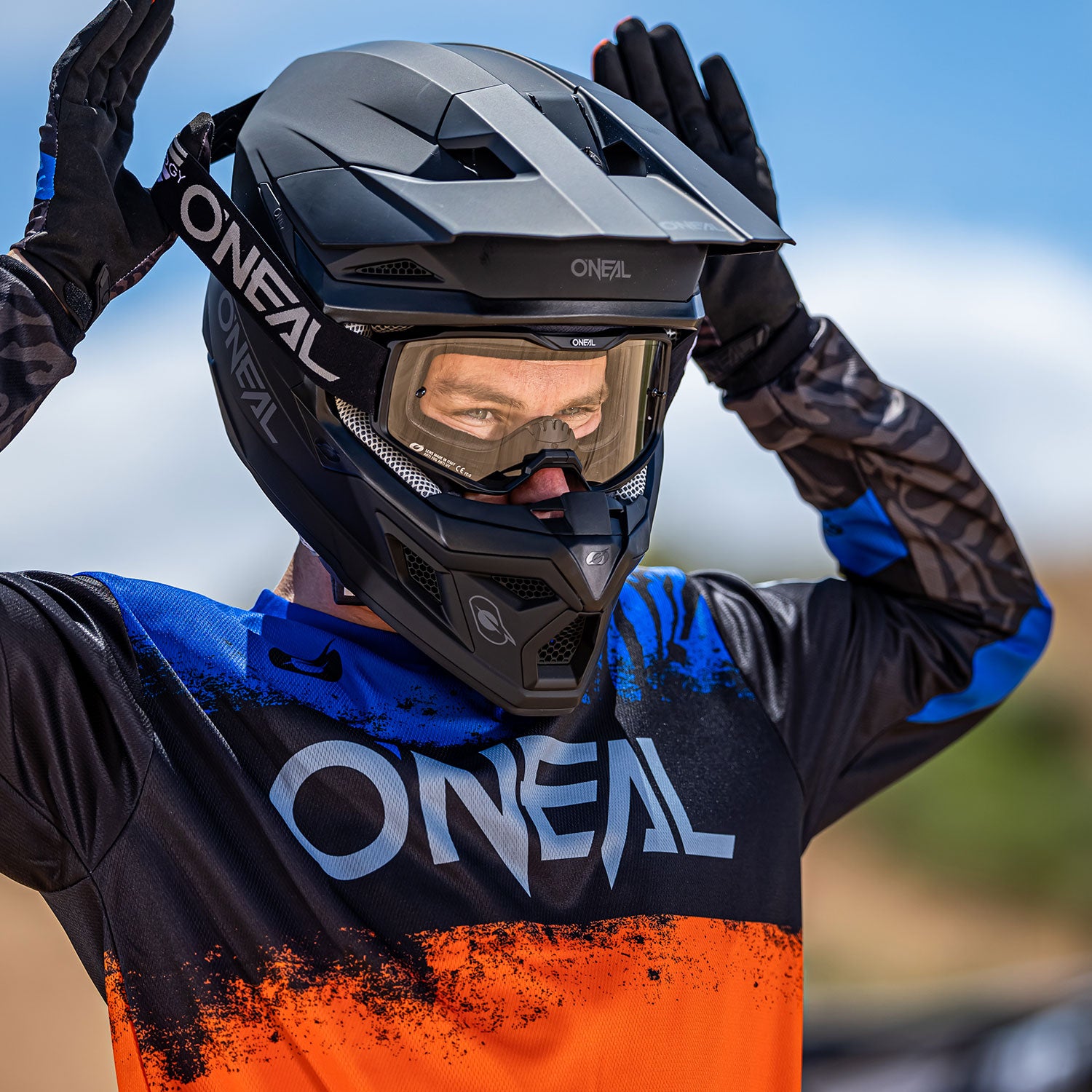 Eine Person in einem schwarz-orange-blauen O'Neal-Motocrosstrikot und einem schwarzen O'Neal-Motocrosshelm mit Schutzbrille steht im Freien. Die Person hat ihre behandschuhten Hände nahe der Oberseite des Helms vor einem blauen Himmelshintergrund erhoben.