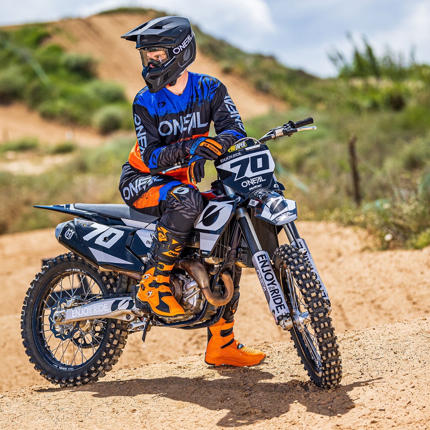 Ein Motocrossfahrer in orange-blauem Rennanzug und Helm sitzt auf einem schwarz-weißen Dirtbike mit der Aufschrift „70“ und „ENJOY RIDE“ auf dem Vorderrad. Der Fahrer ist auf einem Feldweg mit einer natürlichen, grasbewachsenen Landschaft und einem bewölkten Himmel im Hintergrund.