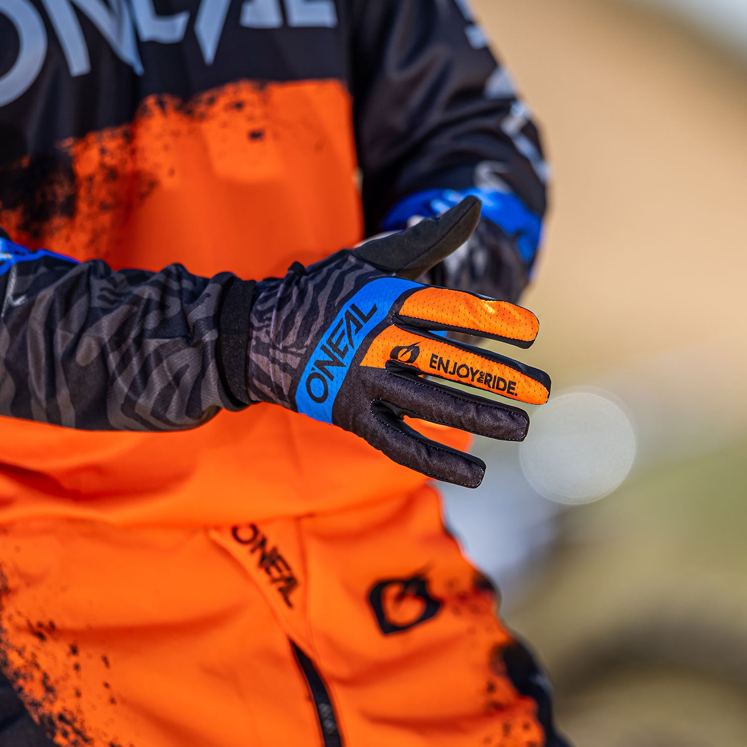 Eine Person in einem orange-schwarzen Outfit mit blauen Akzenten zieht schwarze und orange Handschuhe an, auf denen die Worte „O’NEAL“ und „ENJOYRIDE“ aufgedruckt sind. Der Hintergrund ist unscharf, wodurch die Aufmerksamkeit auf die Handschuhe und das Outfit gelenkt wird.