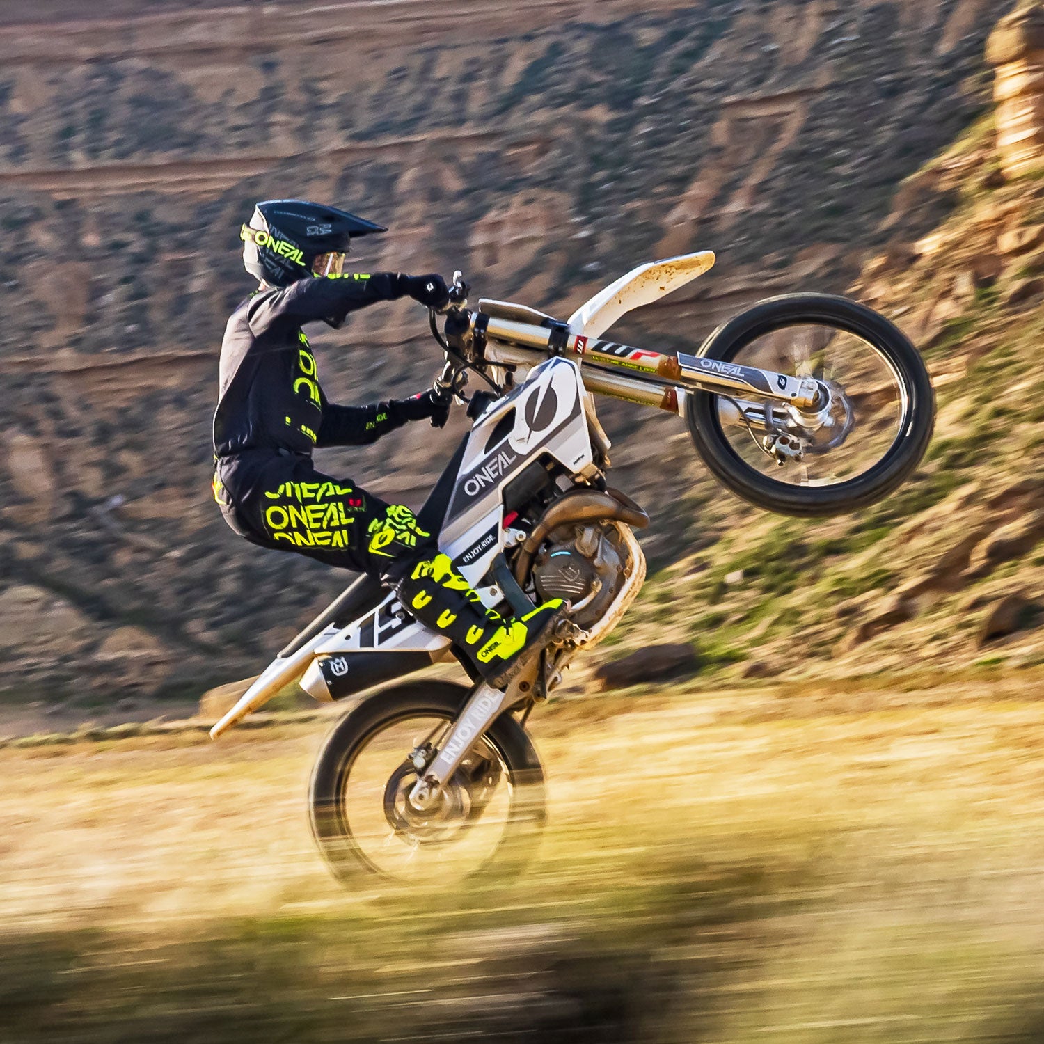 Eine Person in schwarz-neongelber Motocross-Ausrüstung macht auf einem weißen Dirtbike vor einem Hintergrund aus felsigen Hügeln und verschwommenem Vordergrund einen Wheelie, was Geschwindigkeit und Bewegung suggeriert. Der Körper des Fahrers ist nach hinten geneigt und balanciert auf dem Hinterrad des Motorrads.