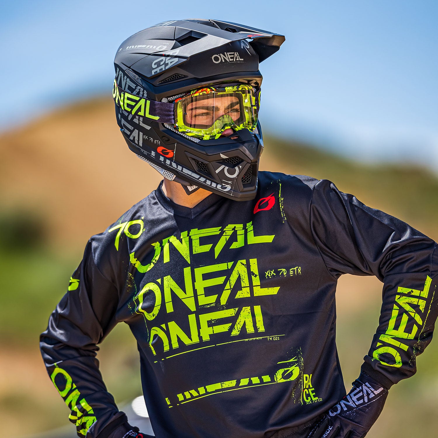 Eine Person, die einen Integralhelm mit Schutzbrille und ein Motocross-Trikot trägt, beide mit der Aufschrift „Oneal“, steht mit ausgestreckten Armen da. Das Trikot hat einen schwarzen Grund mit leuchtend gelbgrüner Beschriftung. Im Hintergrund ist eine verschwommene Landschaft mit Hügeln zu sehen.