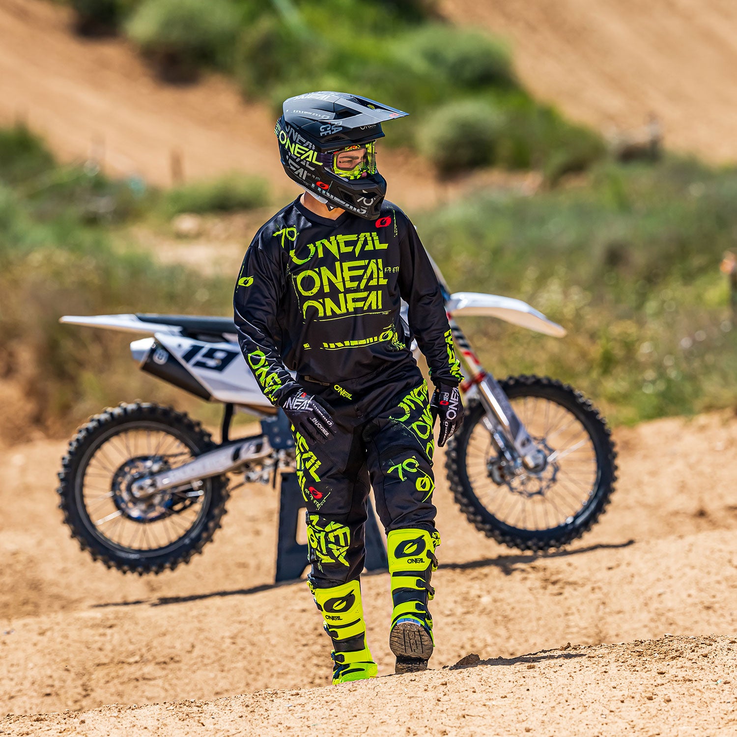 Ein Motocross-Fahrer in schwarz-gelber O'Neal-Ausrüstung und mit Helm steht auf einer unbefestigten Strecke, im Hintergrund ist ein weißes Motocross-Motorrad geparkt.