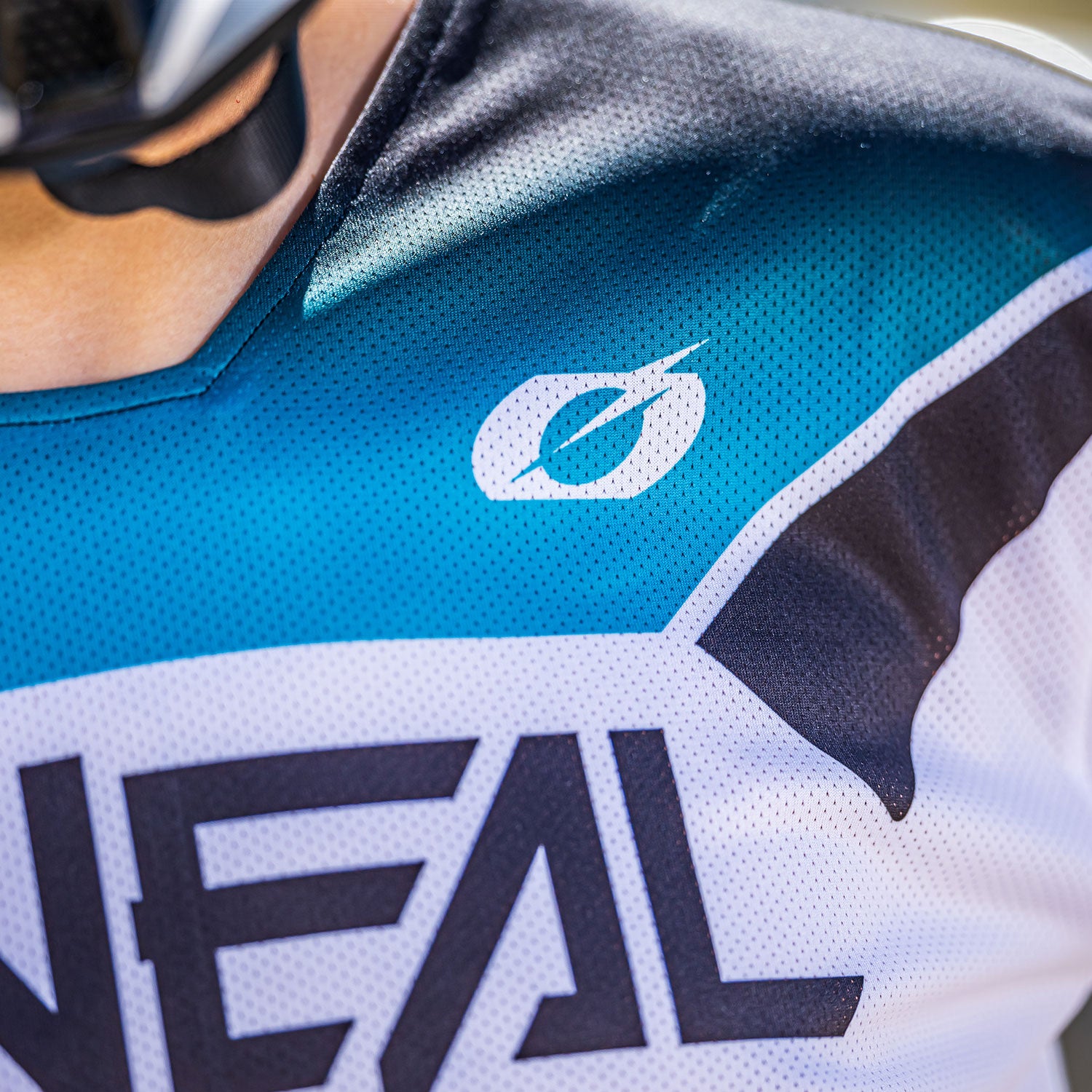 Nahaufnahme einer Person, die ein blau-weißes Sporttrikot mit dem Markennamen „O'NEAL“ und einem stilisierten „O“-Logo auf der Brust trägt. Die Person trägt einen Helm, der nur an seinem Riemen und einem kleinen Teil in der Nähe der linken Schulter zu erkennen ist.