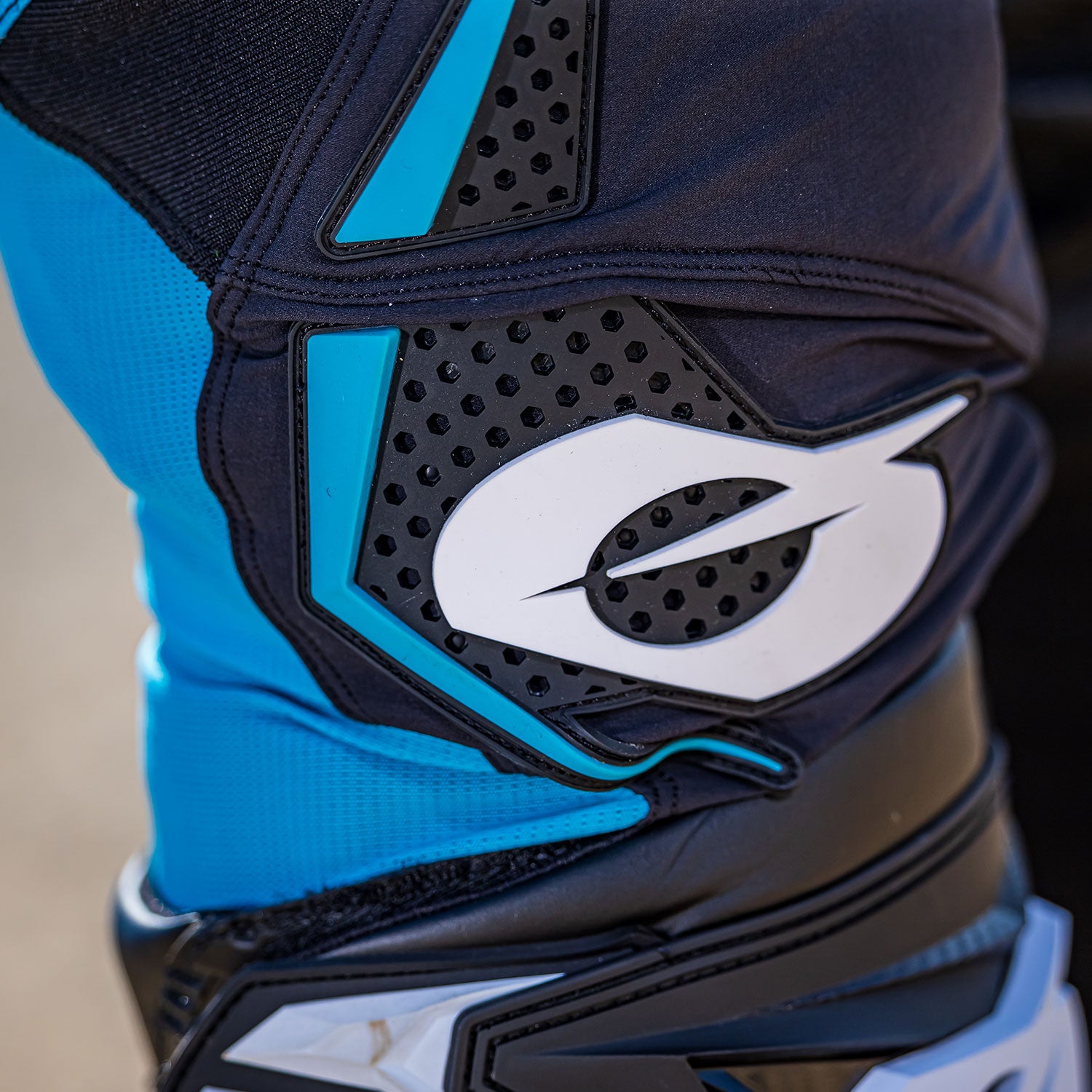 Nahaufnahme eines Knieschoners mit blauen, schwarzen und weißen Details, einschließlich eines markanten weißen Logos, das einem stilisierten Buchstaben „O“ ähnelt. Das Stoffmaterial ist zusammengenäht, wobei schwarze Schutzpolster und blaue Netzabschnitte sichtbar sind.