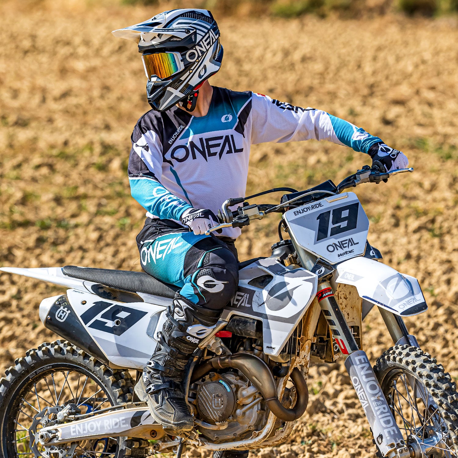 Ein Motocrossfahrer in voller Schutzausrüstung und mit Helm sitzt auf einem weißen Dirtbike. Der Fahrer blickt nach links, während er auf einer Dirtpiste steht. Das Bike trägt die Nummer 19 und auf dem Outfit des Fahrers ist vor allem der Name „O’Neal“ zu sehen.