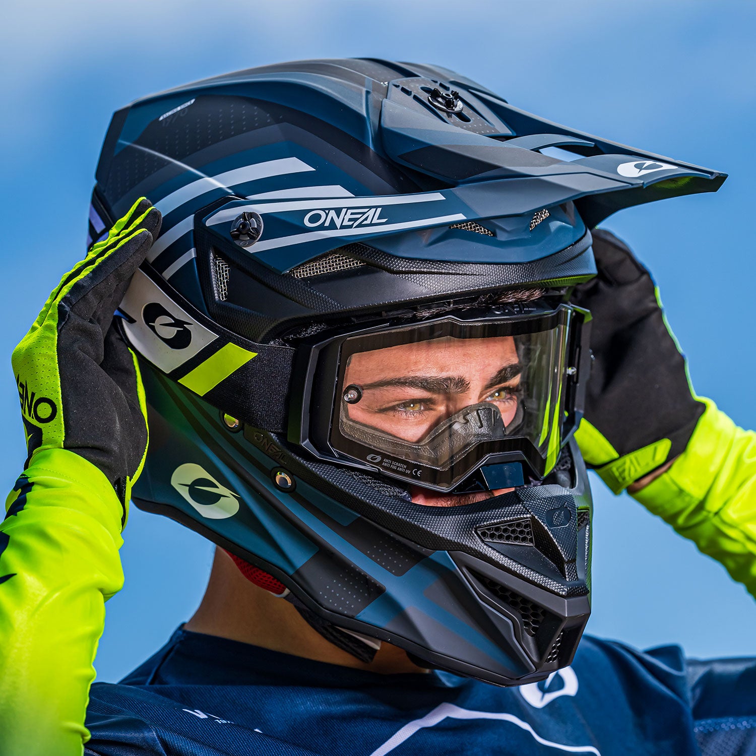 Eine Person mit einem schwarz-blauen Motocross-Helm und einer schwarzen und durchsichtigen Schutzbrille rückt den Helm mit den Händen zurecht. Sie trägt außerdem neongelbe Handschuhe und ein passendes Trikot mit neongelbem Besatz vor einem strahlend blauen Himmelshintergrund.