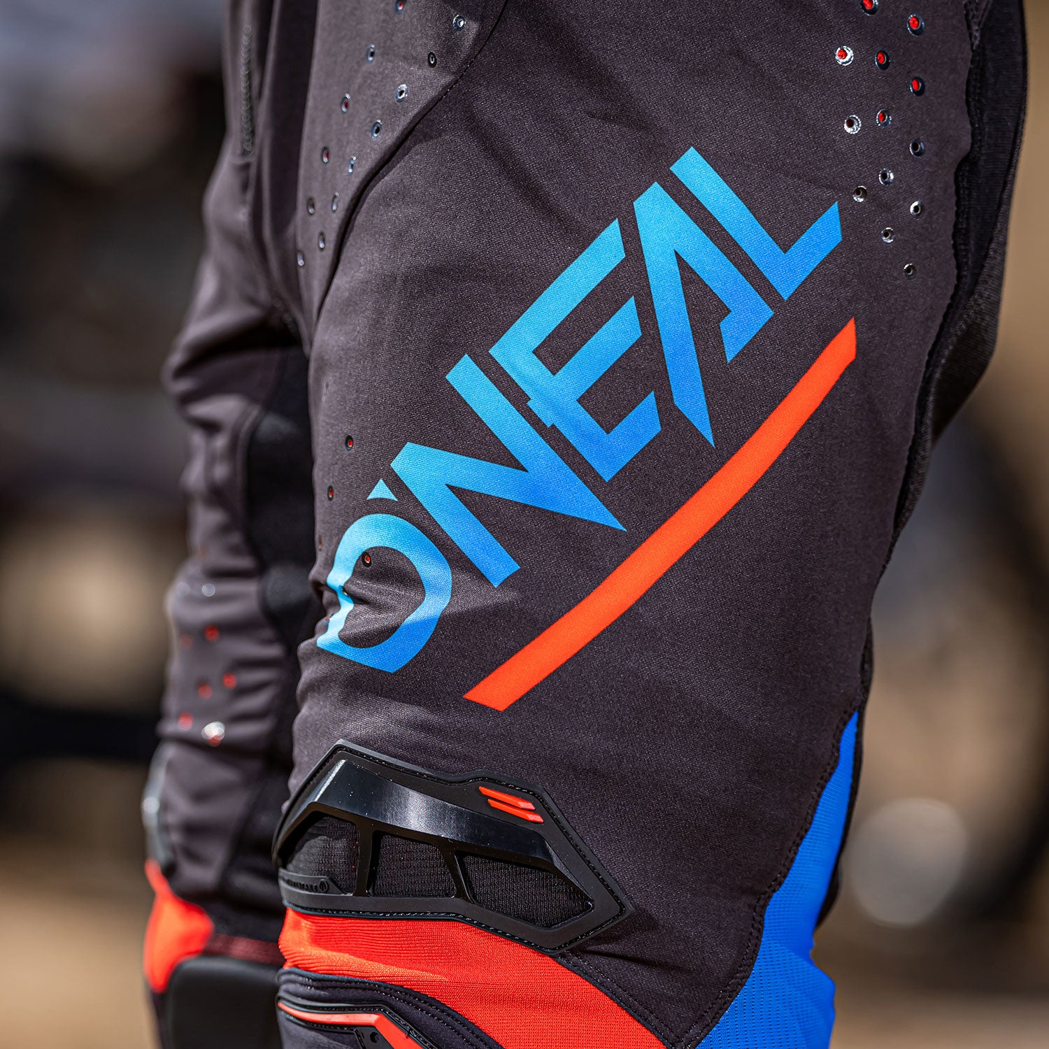 Nahaufnahme einer Person in schwarz-blauer Motocross-Ausrüstung mit dem „O'Neal“-Logo in Blau und Rot, das deutlich auf dem Ärmel zu sehen ist. Die Ausrüstung ist mit Belüftungslöchern und einer schwarzen Schutzpolsterung versehen.