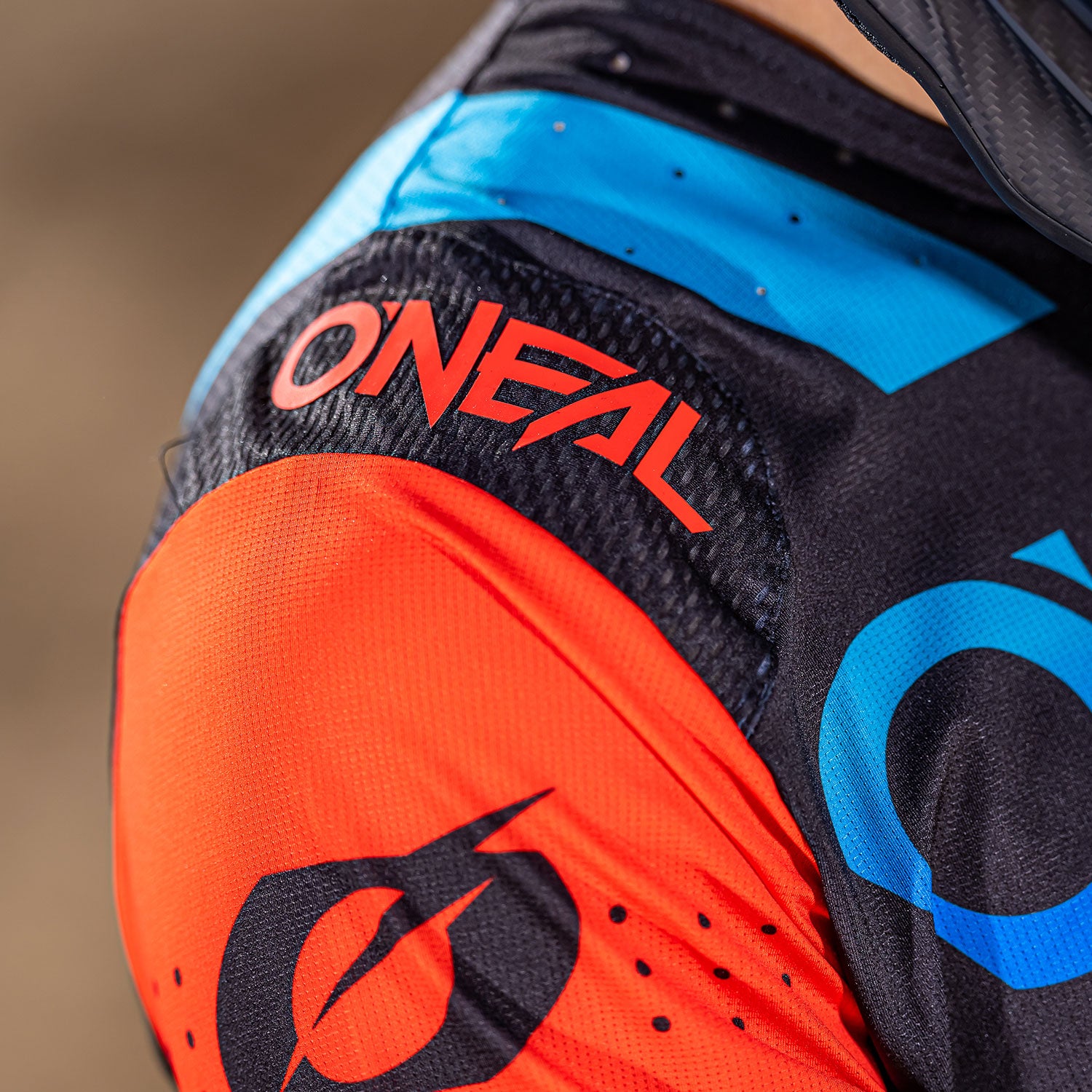Nahaufnahme einer Person, die ein buntes Trikot mit „ONEAL“ und einem stilisierten „O“-Logo trägt. Das Trikot weist Schattierungen von Orange, Schwarz und Blau auf und die sichtbaren Materialien scheinen Hochleistungssportstoffe zu sein.