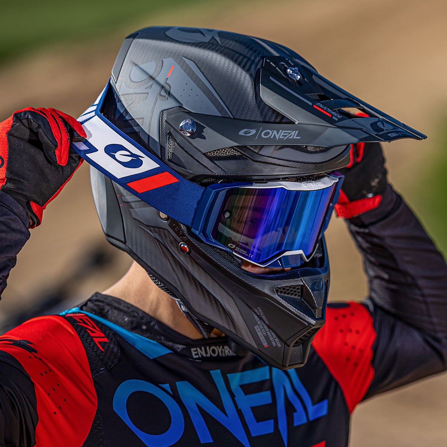 Eine Person in einem schwarz-rot-blauen Motocross-Outfit und einem schwarzen Helm mit getöntem Visier rückt ihre Schutzbrille zurecht. Helm und Ausrüstung sind mit dem Logo „Oneal“ versehen. Im Hintergrund ist eine Dirt-Track-Strecke im Freien zu sehen.