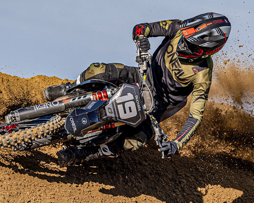 Ein Motocross-Fahrer in schwarzer und grüner Montur lehnt sich auf einer unbefestigten Strecke an und wirbelt dabei Erde auf, während er ein schwarzes Dirt Bike mit der Nummer 19 fährt. Der Fahrer trägt einen Helm und Handschuhe, der Himmel ist im Hintergrund zu sehen.