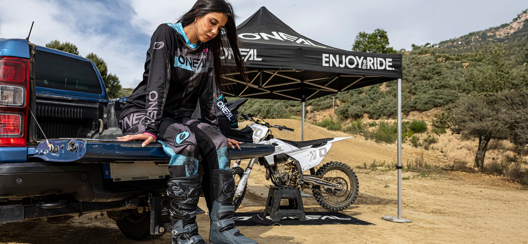Eine Frau in Motocross-Ausrüstung sitzt auf der Heckklappe eines blauen Pickups in der Nähe eines Dirt-Bikes und eines schwarzen Zelts mit der Aufschrift ENJOY RIDE", im Hintergrund eine hügelige Landschaft.
