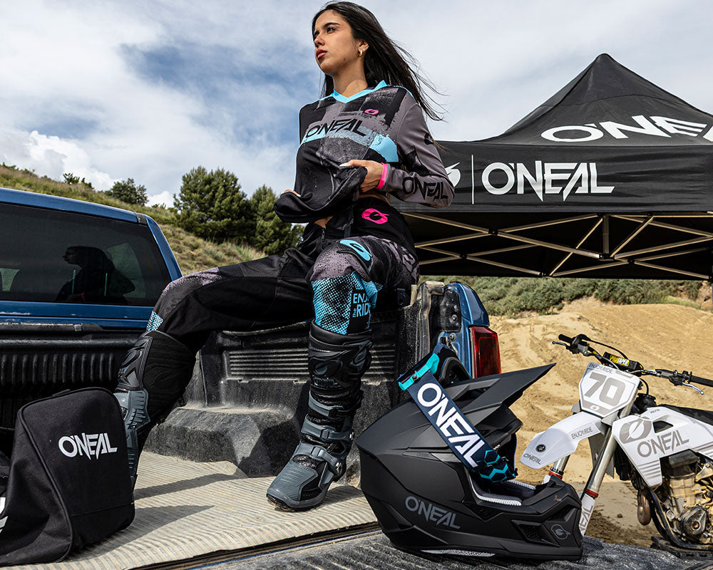 Eine Frau in Motocross-Kleidung sitzt mit Handschuhen auf der Ladefläche eines Lastwagens, in der Nähe ein Helm. Ein kleines Dirt Bike und ein Zelt der Marke O'Neal sind im Hintergrund zu sehen.
