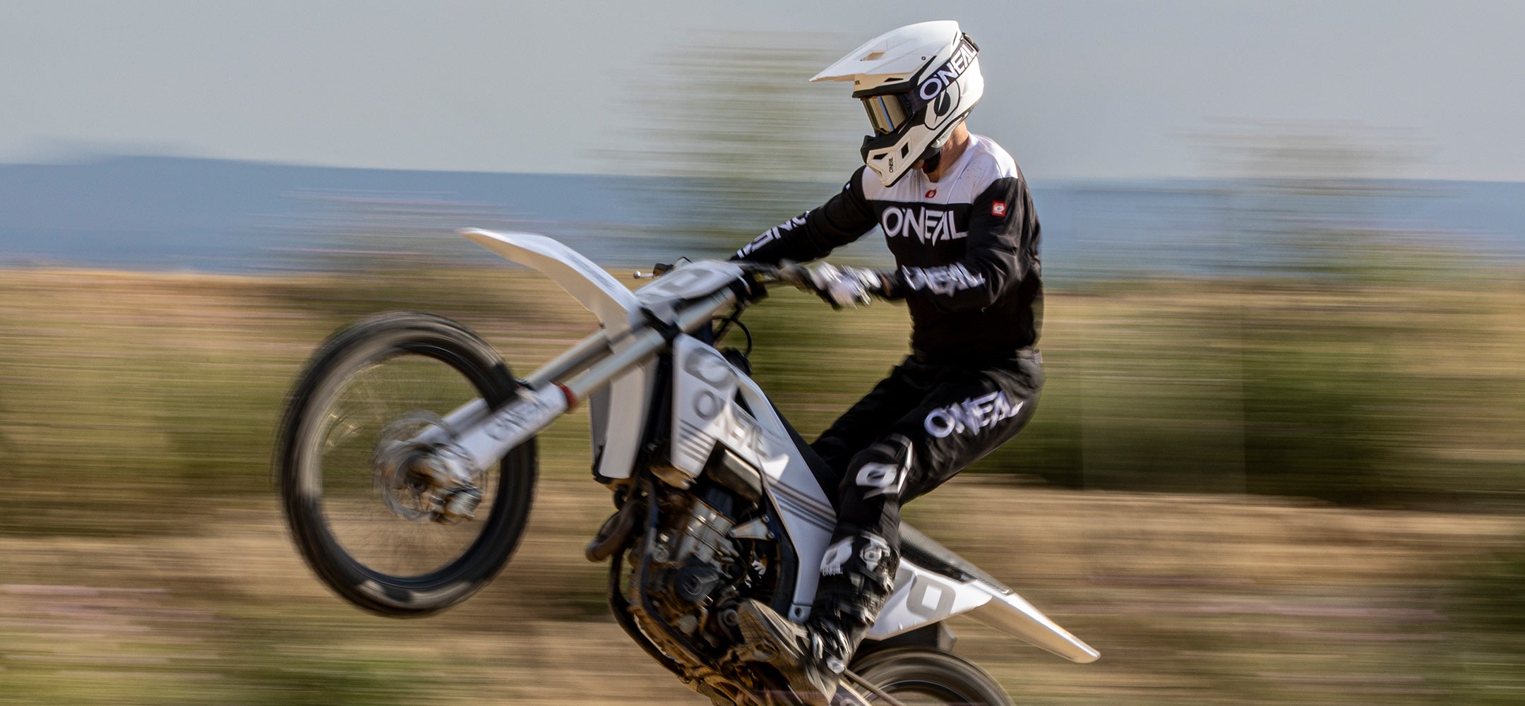 Ein Motocross-Fahrer in schwarzer und weißer Ausrüstung und Helm vollführt einen Wheelie auf einem Dirt Bike, wobei der verschwommene Hintergrund eine schnelle Bewegung im Freien suggeriert.