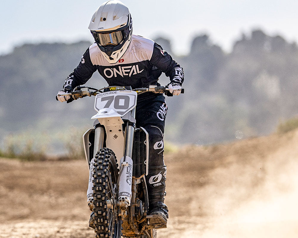 Ein Motocross-Fahrer mit weißem Helm und schwarzer O'Neal-Kleidung fährt auf einem Dirt Bike mit der Nummer 70 auf einer staubigen Outdoor-Strecke, mit unscharfen Bäumen im Hintergrund.
