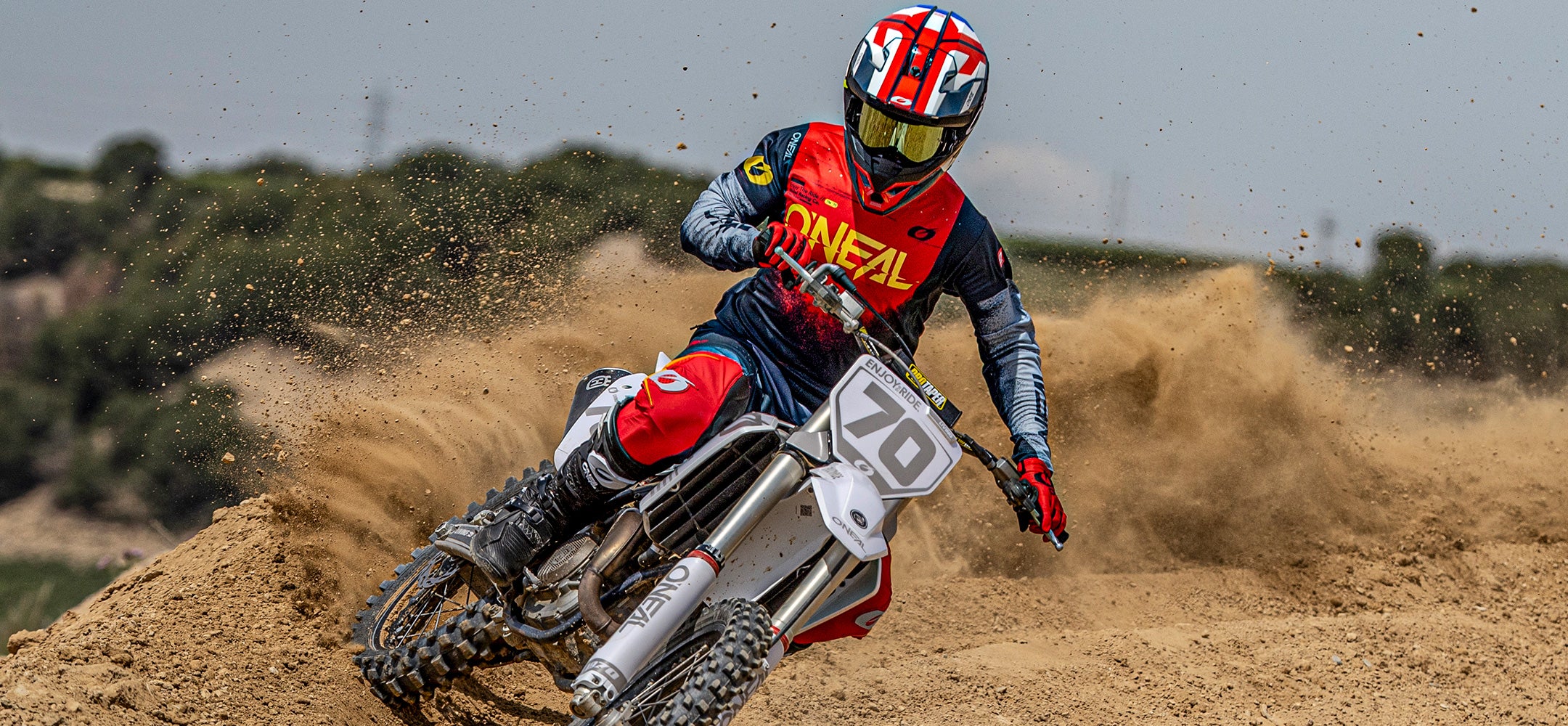 Ein Motocross-Fahrer in roter, blauer und gelber Kleidung rast auf einem weißen Dirt Bike um eine sandige Kurve und wirbelt dabei eine große Staubwolke auf. Der Fahrer trägt zum Schutz einen Helm und eine Schutzbrille.