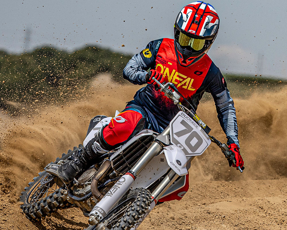 Ein Motocross-Fahrer in roter und blauer O'Neal-Kleidung rast auf einer unbefestigten Strecke und wirbelt Staub auf, während er auf einem weißen Motorrad mit der Nummer 70 fährt. Der Fahrer trägt einen roten Helm mit Schutzbrille.