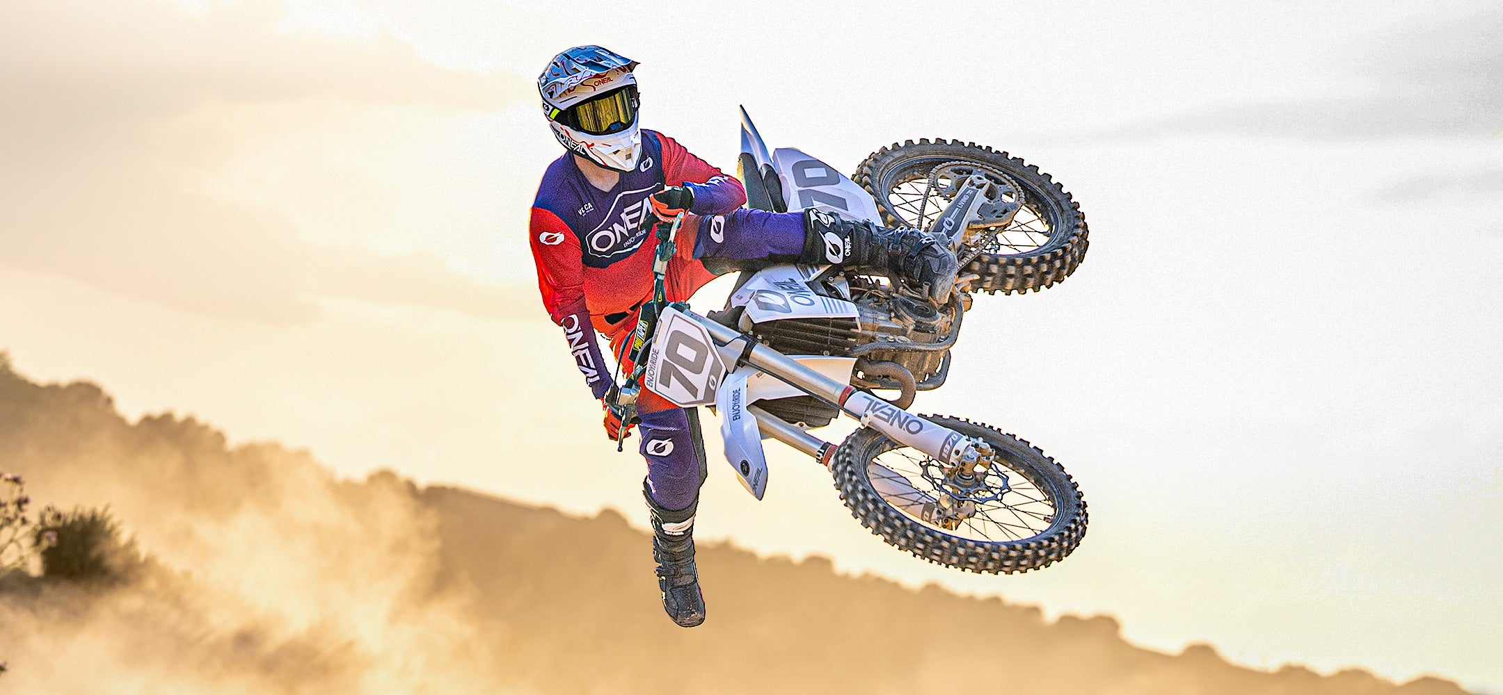 Ein Motocross-Fahrer in roter und blauer Montur vollführt einen Trick in der Luft auf einem Dirt Bike, während unter den Rädern Staub aufsteigt und im Hintergrund ein heller, dunstiger Himmel zu sehen ist.