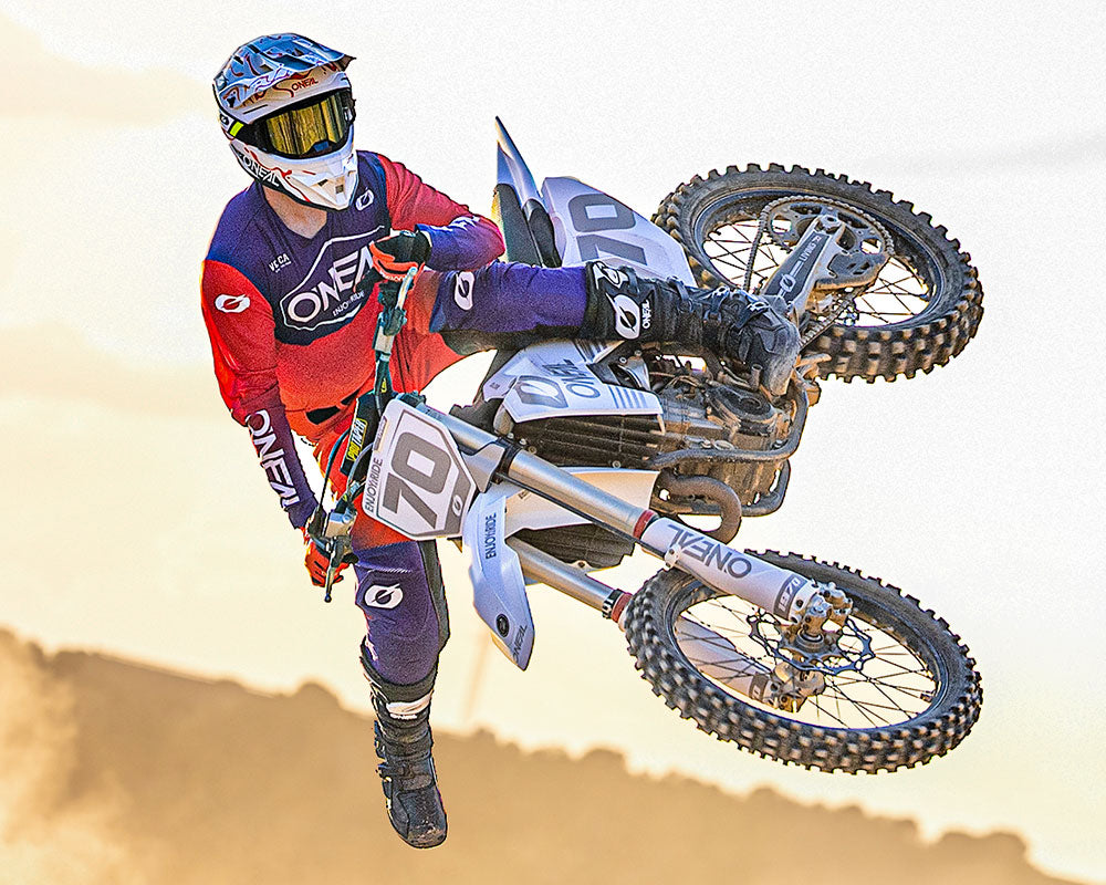 Ein Motocross-Fahrer in farbenfroher Ausrüstung und mit Helm vollführt einen Stunt in der Luft auf einem Dirt Bike, wobei der Hintergrund unscharf und staubig ist, was auf Geschwindigkeit und Action hindeutet.