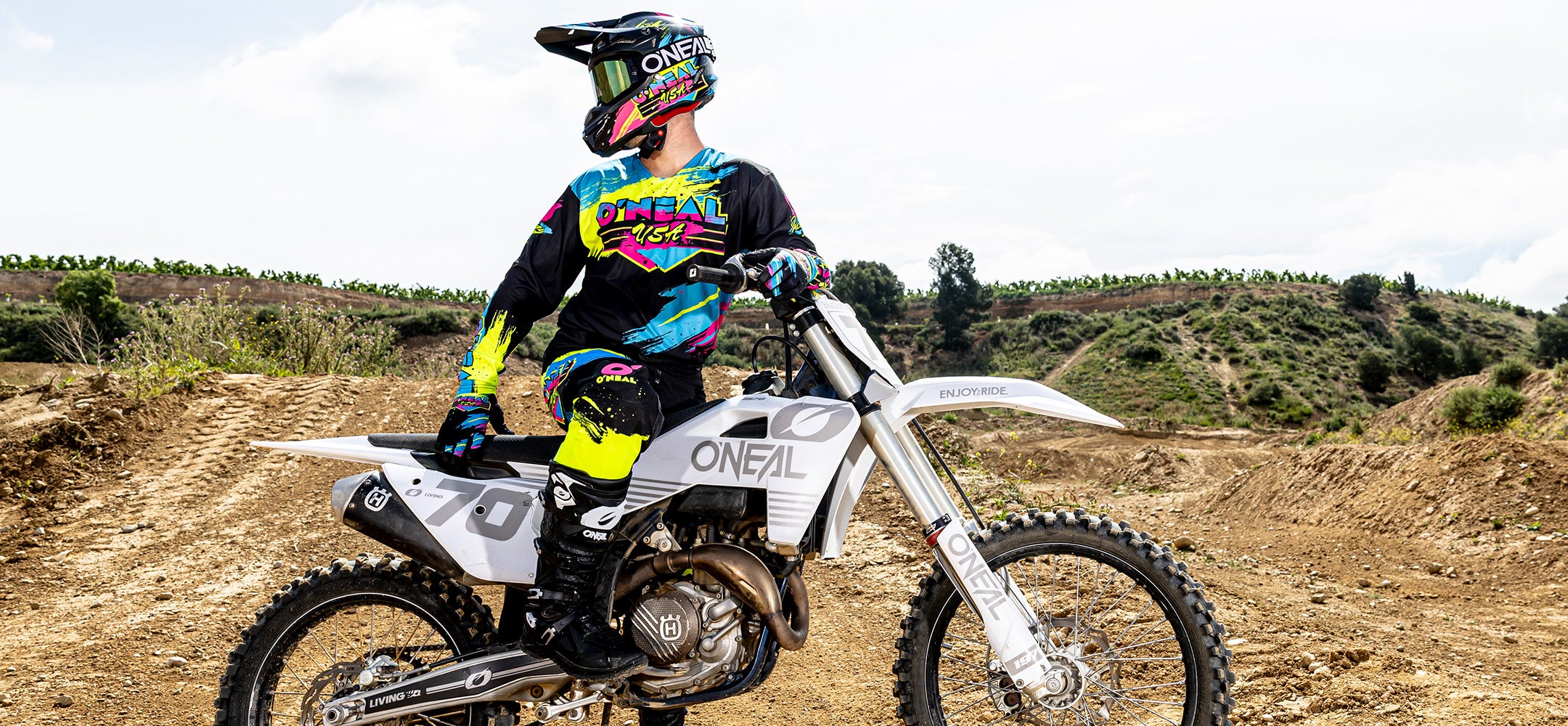 Ein Motocross-Fahrer in farbenfroher O'Neal-Kleidung und Helm sitzt auf einem weißen Dirt Bike, das auf einer unbefestigten Strecke mit Hügeln und Grünflächen im Hintergrund geparkt ist. Der Fahrer blickt zur Seite, die Hände auf dem Lenker.