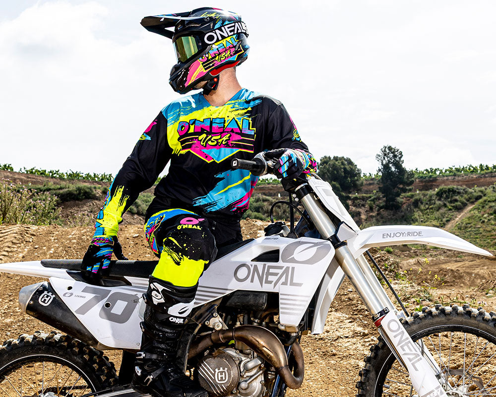 Ein Motocross-Fahrer in einem farbenfrohen O'Neal-Anzug und einem Helm sitzt auf einem weißen Dirt Bike mit der Aufschrift "70" und "ONEAL" auf einer unbefestigten Strecke mit Grünzeug und einem bewölkten Himmel im Hintergrund.