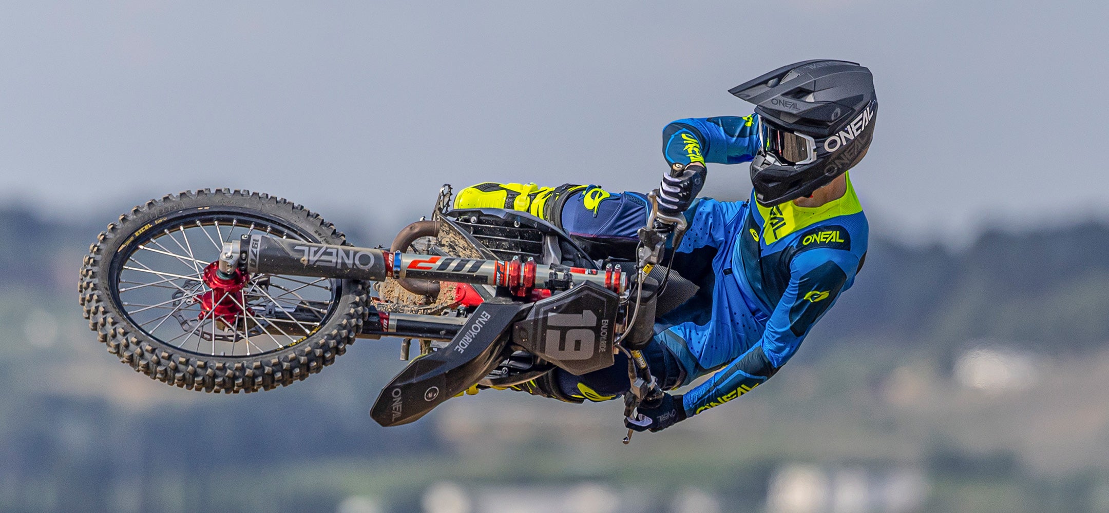 Ein Motocross-Fahrer in einem blau-gelben Anzug und mit schwarzem Helm führt einen Sprung auf einem Dirt Bike aus, wobei das Motorrad seitlich in der Luft vor einem unscharfen Hintergrund im Freien gekippt wird.